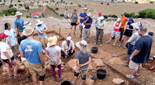 People at an archaeological dig stand and listen to someone squatting down in the work area